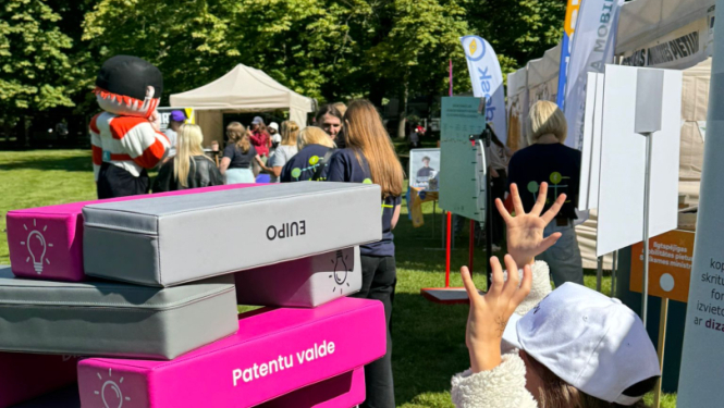 Meitene ar baltu cepuri spēlē lielizmēra Jenga torni ar pelēkiem un rozā klucīšiem, uz kuriem rakstīts “Patentu valde”.