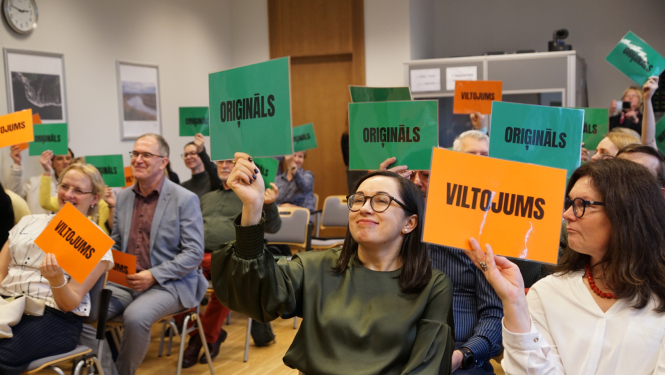 semināra dalībnieki rāda planšetes ar uzrakstiem oriģināls un vltojums piedaloties spēlē un mēģinot atpazīt rādītos piemērus