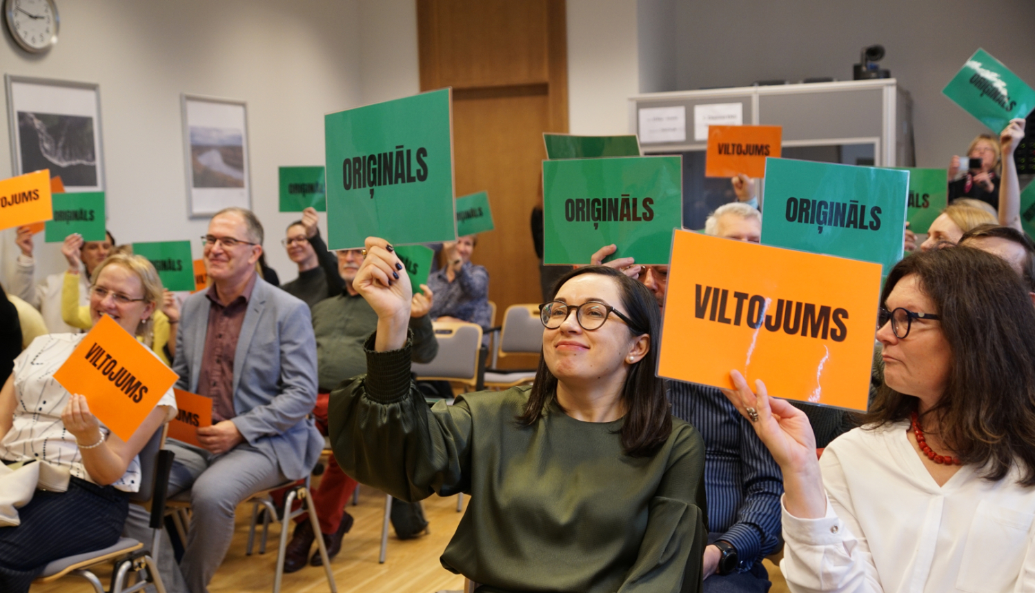 semināra dalībnieki rāda planšetes ar uzrakstiem oriģināls un vltojums piedaloties spēlē un mēģinot atpazīt rādītos piemērus