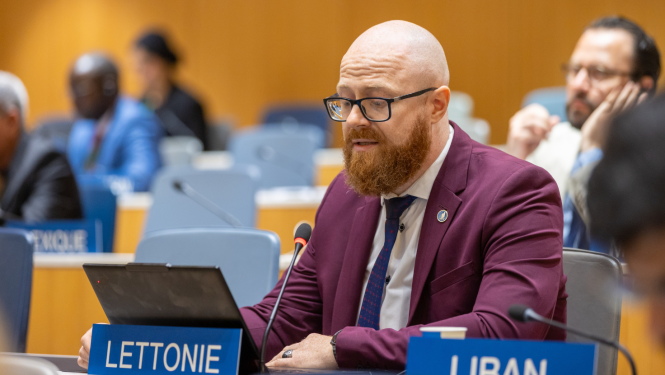 Patentu valdes direktors sēž konferenču telpā pie galda, Uz galda atrodas dators un miikrafons statīvā. Fonāredzama konferenču telpa ar pārējiem dalībniekiem, priekšā novietota plāksnītē ar uzrakstu Latvija.