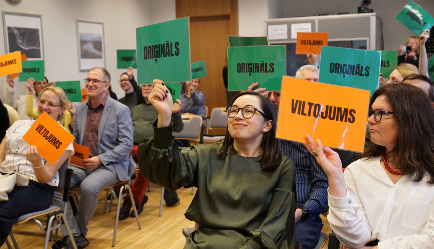 semināra dalībnieki rāda planšetes ar uzrakstiem oriģināls un vltojums piedaloties spēlē un mēģinot atpazīt rādītos piemērus